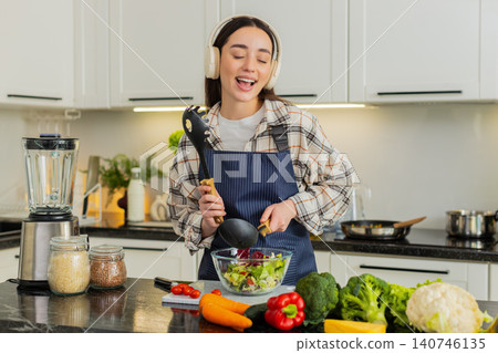 Young woman dancing with kitchen utensils while making keto veggie salad at home apartment indoors Young woman dancing with kitchen utensils while making keto veggie salad at home apartment indoors 140746135