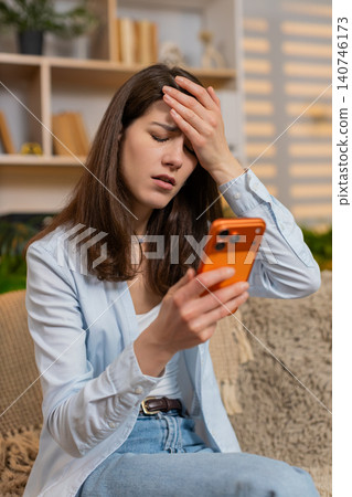 Young woman on home sofa reading bad news on smartphone showing shock stress upset reaction indoors 140746173