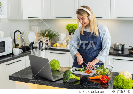 Young woman cutting cucumber for keto salad while watching recipe on laptop and wearing headphones 140746208