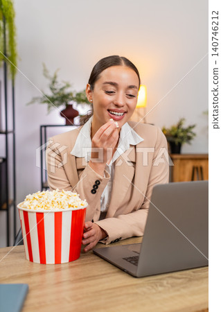 Young businesswoman enjoys popcorn during break launches series trailer on laptop and giggles today 140746212
