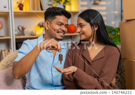 Indian couple man and woman sitting on home sofa showing new home keys and celebrating housing joy Indian couple man and woman sitting on home sofa showing new home keys and celebrating housing joy 140746222