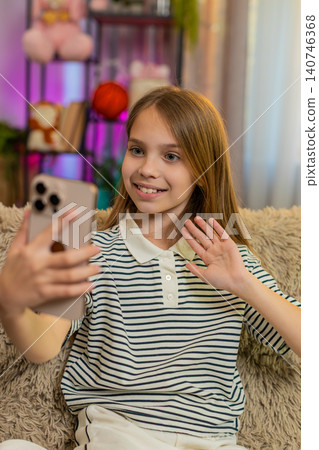 Child girl kid smiles and waves during video call on smartphone, talks happily sitting alone on sofa Child girl kid smiles and waves during video call on smartphone, talks happily sitting alone on sofa 140746368