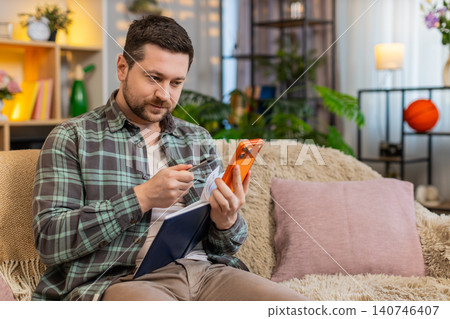 Young man on home sofa using smartphone checking task list and writing new idea in notebook at home Young man on home sofa using smartphone checking task list and writing new idea in notebook at home 140746407