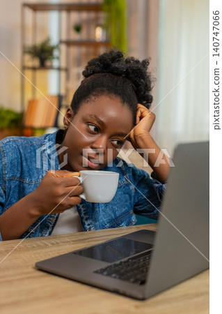 African American young woman drinks coffee tiredly before remote work on laptop sitting at home African American young woman drinks coffee tiredly before remote work on laptop sitting at home 140747066