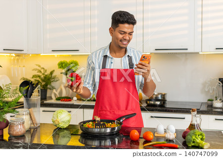 Indian man frying vegetables while checking grocery list on smartphone for healthy meal prep at home 140747099