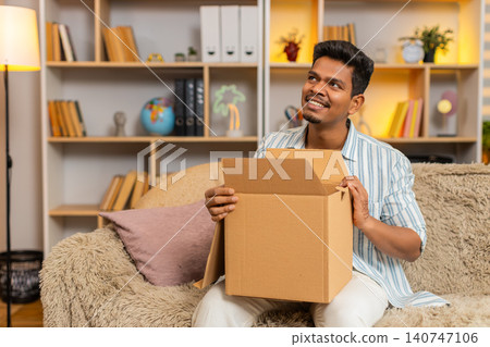 Indian man on home sofa unboxing delivery box with surprise and joy after online shopping purchase Indian man on home sofa unboxing delivery box with surprise and joy after online shopping purchase 140747106