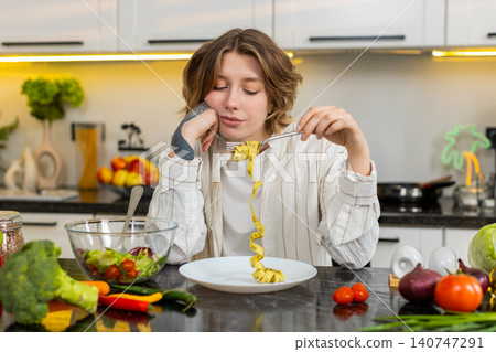 Young woman feeling upset by diet salad in kitchen while holding tape measure and losing motivation Young woman feeling upset by diet salad in kitchen while holding tape measure and losing motivation 140747291