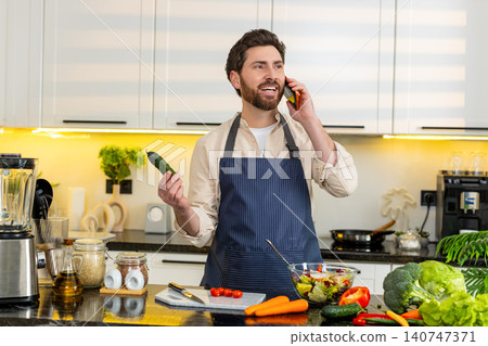Middle-aged man tasting fresh vegetable salad while talking on smartphone in home kitchen for diet 140747371
