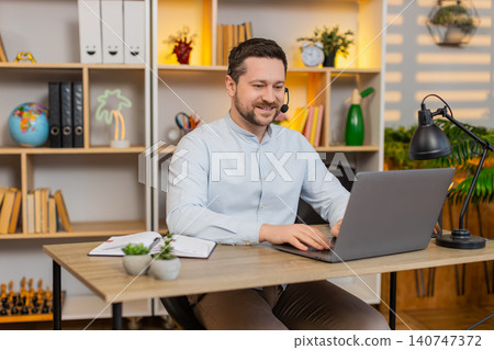 Young man on home table working in headset with laptop and smiling during virtual customer support Young man on home table working in headset with laptop and smiling during virtual customer support 140747372