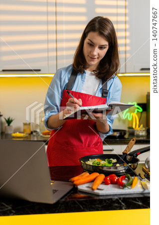 Young woman taking notes from laptop during cooking class for healthy diet meal planning indoor home Young woman taking notes from laptop during cooking class for healthy diet meal planning indoor home 140747667