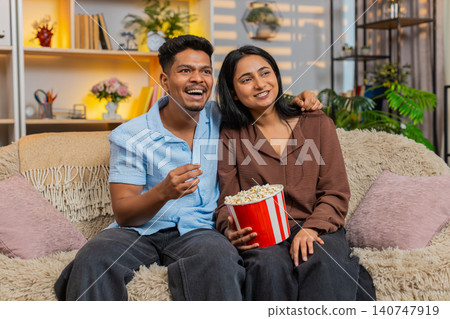 Indian couple man and woman sitting on home sofa watching comedy movie with popcorn in home leisure 140747919