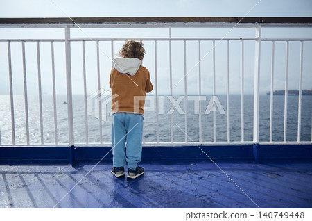 Young boy stands at a ferry railing, gazing across calm open sea toward the distant horizon. The scene suggests travel, solitude, childhood curiosity and a peaceful coastal journey on a bright day. 140749448