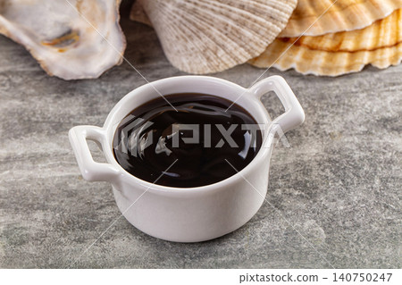 White ceramic bowl filled with dark sauce beside seashells on textured surface White ceramic bowl filled with dark sauce beside seashells on textured surface 140750247