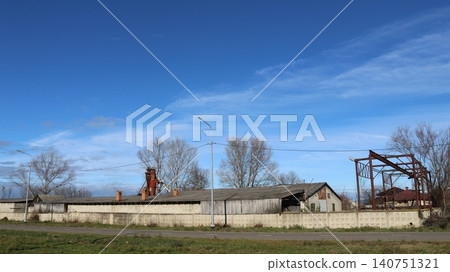 A long, one-story building, reminiscent of an old hangar, warehouse, or cowshed. To the left, a tall, rusty industrial structure resembling a grain cleaning complex or elevator is visible. A long, one-story building, reminiscent of an old hangar, warehouse, or cowshed. To the left, a tall, rusty industrial structure resembling a grain cleaning complex or elevator is visible. 140751321