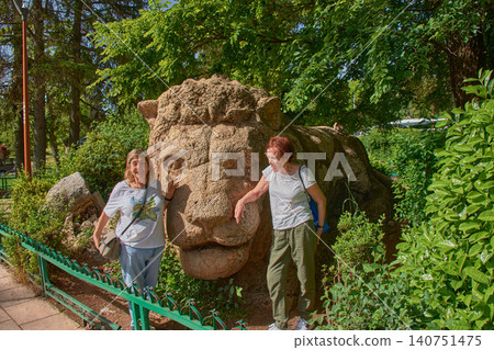 Two female tourists smile happily, posing next to the famous stone sculpture of the Lion of Ifrane in Morocco. Two female tourists smile happily, posing next to the famous stone sculpture of the Lion of Ifrane in Morocco. 140751475