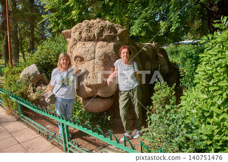 Two female tourists smile happily, posing next to the famous stone sculpture of the Lion of Ifrane in Morocco. Two female tourists smile happily, posing next to the famous stone sculpture of the Lion of Ifrane in Morocco. 140751476