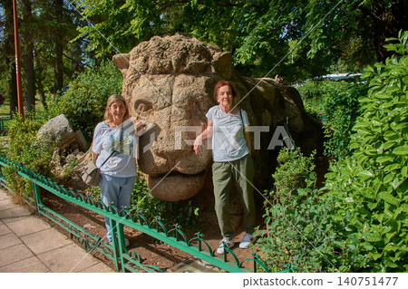 Two female tourists smile happily, posing next to the famous stone sculpture of the Lion of Ifrane in Morocco. 140751477