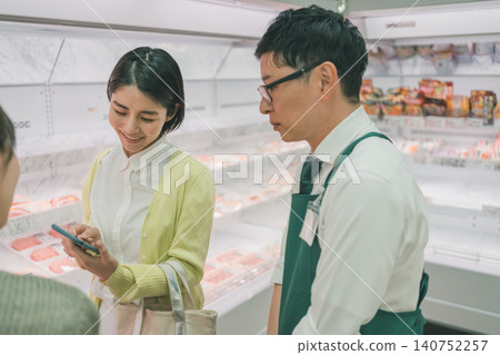 一位女顧客在超市肉類區向一位男店員展示她的智慧型手機並尋求建議。 140752257