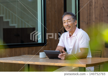一位老年男子在露台上一邊喝咖啡一邊操作平板電腦。 一位老年男子在露台上一邊喝咖啡一邊操作平板電腦。 140752783