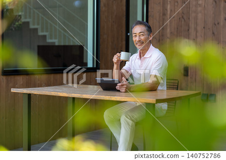 一位老年男子在露台上一邊喝咖啡一邊操作平板電腦。 140752786