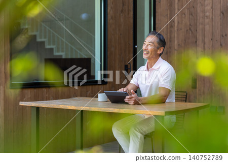一位老年男子在露台上一邊喝咖啡一邊操作平板電腦。 一位老年男子在露台上一邊喝咖啡一邊操作平板電腦。 140752789