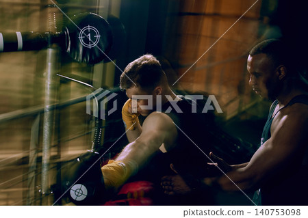 Coach supporting boxer during fast reflex bag workout in dark gym. 140753098