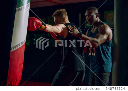 Boxing coach guiding athlete punch technique on heavy bag in dark gym. Boxing coach guiding athlete punch technique on heavy bag in dark gym. 140753124