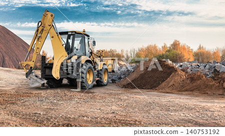 Universal excavator loader operates on a construction site in the daytime. The machine is moving dirt and rocks while surrounded by piles of material Universal excavator loader operates on a construction site in the daytime. The machine is moving dirt and rocks while surrounded by piles of material 140753192