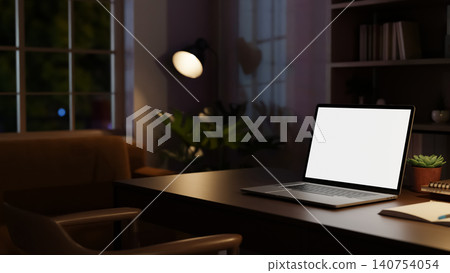 Dark home office with white screen laptop on wooden table beside bookshelf illuminated by floor lamp Dark home office with white screen laptop on wooden table beside bookshelf illuminated by floor lamp 140754054
