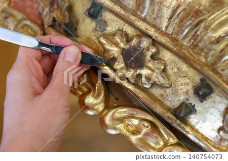 Craftsman Works on Detailed Restoration of a Gilded Wood Sculpture in a Workshop Setting During Daylight Hours 140754073