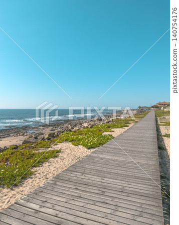 A beautiful long wooden boardwalk stretches alongside the rocky Atlantic shore and sandy dunes of Praia da Granja in Porto Portugal under a bright clear blue sky during a sunny summer day A beautiful long wooden boardwalk stretches alongside the rocky Atlantic shore and sandy dunes of Praia da Granja in Porto Portugal under a bright clear blue sky during a sunny summer day 140754176