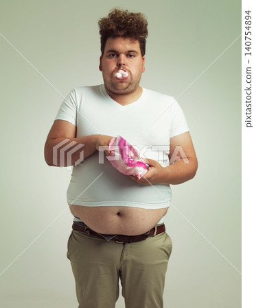 Food, portrait and plus size man with marshmallow, dessert and candy in a studio. Bag, sweet and unhealthy snack for over eating a treat with funny face and hungry male person with grey background Food, portrait and plus size man with marshmallow, dessert and candy in a studio. Bag, sweet and unhealthy snack for over eating a treat with funny face and hungry male person with grey background 140754894