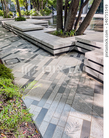 A sophisticated urban building with dappled sunlight filtering through the trees, and a plaza with stone benches. 140755549