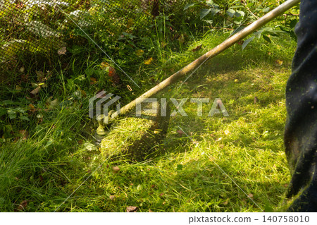 Man professional gardener using grass trimmer to cut lawn in backyard. Manual labor for landscape maintenance. Lawn mower tool for gardening and yard work. Grass cutting agricultural activity. Man professional gardener using grass trimmer to cut lawn in backyard. Manual labor for landscape maintenance. Lawn mower tool for gardening and yard work. Grass cutting agricultural activity. 140758010