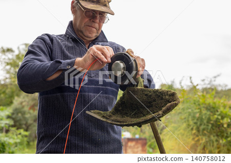 Man changing orange trimmer line on string trimmer head. Gardener reparing power tool for lawn maintenance. Grass mowing equipment service, repair and garden work preparation concept. 140758012