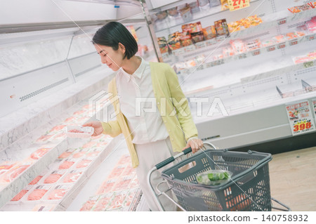 一位女顧客在超市肉品區拿起一包肉仔細查看，旁邊放著一個裝滿蔬菜的購物籃。 140758932