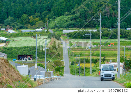 A light van stands on a peaceful country road surrounded by lush mountains and fields. A light van stands on a peaceful country road surrounded by lush mountains and fields. 140760182