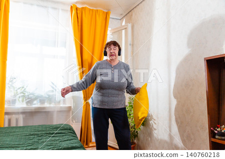 Caucasian senior woman dancing joyfully while wearing headphones at home. Wellness and healthy lifestyle in retirement. Mid shot Caucasian senior woman dancing joyfully while wearing headphones at home. Wellness and healthy lifestyle in retirement. Mid shot 140760218