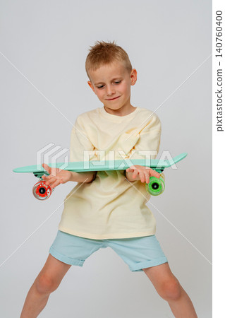 Dynamic full length portrait of a playful young boy posing with a mint green skateboard turned sideways. Bright studio background, showcasing happy emotions, children's style, and sports. Cheerful Dynamic full length portrait of a playful young boy posing with a mint green skateboard turned sideways. Bright studio background, showcasing happy emotions, children's style, and sports. Cheerful 140760400