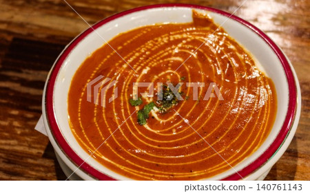 close up of Indian butter chicken curry with cream swirl and cilantro herb garnish with cream swirl on top in a white bowl with red rim on wooden table and close space close up of Indian butter chicken curry with cream swirl and cilantro herb garnish with cream swirl on top in a white bowl with red rim on wooden table and close space 140761143