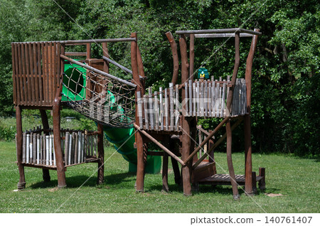 Wooden playground structure with slide and climbing net in grassy park setting Wooden playground structure with slide and climbing net in grassy park setting 140761407