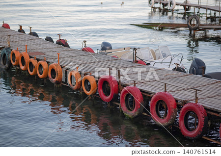 Wooden dock with colorful buoy rings and boats moored on calm water surface Wooden dock with colorful buoy rings and boats moored on calm water surface 140761514