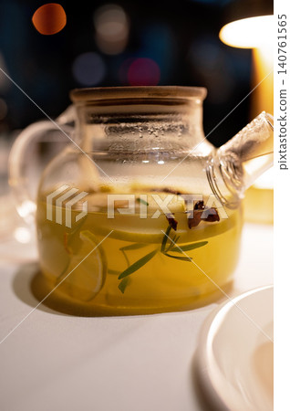 Glass teapot with herbal infusion containing lemon slices and herbs on a table with soft lighting 140761565