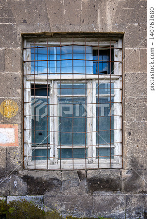 Weathered stone building with barred, broken window showing sky reflection Weathered stone building with barred, broken window showing sky reflection 140761680