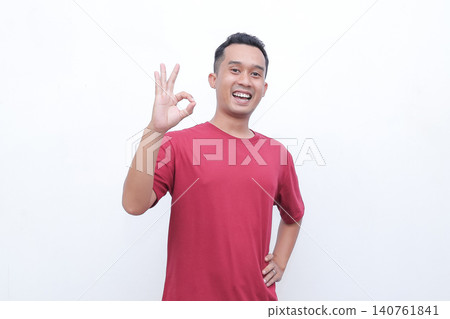 Young man wears red tshirt. He smiles broadly with joy and enthusiasm. Both hands form OK gestures, showing approval. Simple white background enhances focus on his expression Young man wears red tshirt. He smiles broadly with joy and enthusiasm. Both hands form OK gestures, showing approval. Simple white background enhances focus on his expression 140761841
