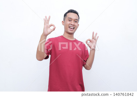 Young man wears red tshirt. He smiles broadly with joy and enthusiasm. Both hands form OK gestures, showing approval. Simple white background enhances focus on his expression Young man wears red tshirt. He smiles broadly with joy and enthusiasm. Both hands form OK gestures, showing approval. Simple white background enhances focus on his expression 140761842
