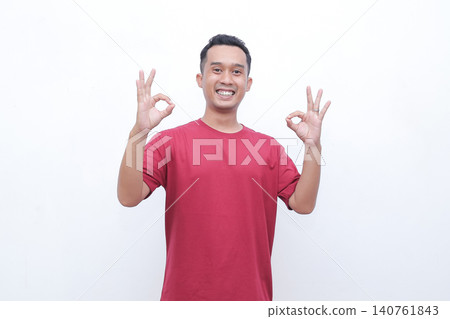 Young man wears red tshirt. He smiles broadly with joy and enthusiasm. Both hands form OK gestures, showing approval. Simple white background enhances focus on his expression Young man wears red tshirt. He smiles broadly with joy and enthusiasm. Both hands form OK gestures, showing approval. Simple white background enhances focus on his expression 140761843