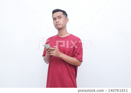 Young man in red shirt holds smartphone. He looks thoughtful, focused on screen. Simple background emphasizes his expression. Ideal for tech, business, or daily life concepts Young man in red shirt holds smartphone. He looks thoughtful, focused on screen. Simple background emphasizes his expression. Ideal for tech, business, or daily life concepts 140761851