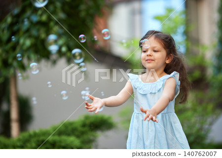 Young girl in a light blue dress with long hair joyfully blowing soap bubbles in a sunny green garden, carefree summer playtime Young girl in a light blue dress with long hair joyfully blowing soap bubbles in a sunny green garden, carefree summer playtime 140762007