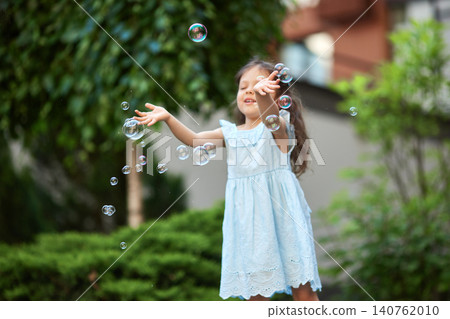 Happy child in a blue dress playing with soap bubbles in a green garden, experiencing joy and carefree childhood moments Happy child in a blue dress playing with soap bubbles in a green garden, experiencing joy and carefree childhood moments 140762010